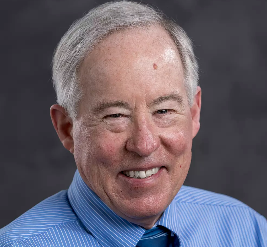 An older man with short gray hair smiles at the camera. He is wearing a blue striped shirt and matching tie, set against a dark, blurred background.
